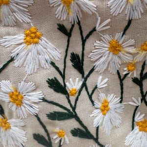 May include: Close-up of a beige fabric embroidered with white daisies with yellow centers and green stems and leaves. The floral embroidery is detailed and textured, creating a natural, handcrafted look.