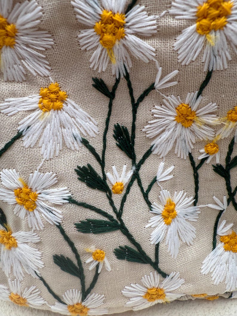May include: Close-up of a beige fabric embroidered with white daisies with yellow centers and green stems and leaves. The floral embroidery is detailed and textured, creating a natural, handcrafted look.