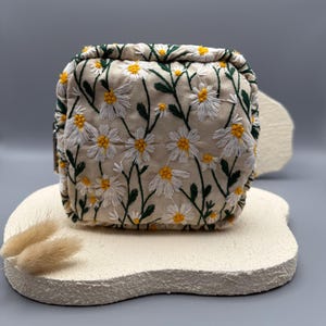May include: A beige, square cosmetic bag with a daisy floral embroidery design. The bag features white daisies with yellow centers and green stems and leaves. The bag is sitting on a textured, off-white surface.