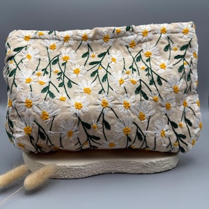 May include: A beige clutch with an embroidered daisy pattern. The daisies have white petals and yellow centers, with green stems and leaves. The clutch is rectangular and sits on a light-colored, textured surface.