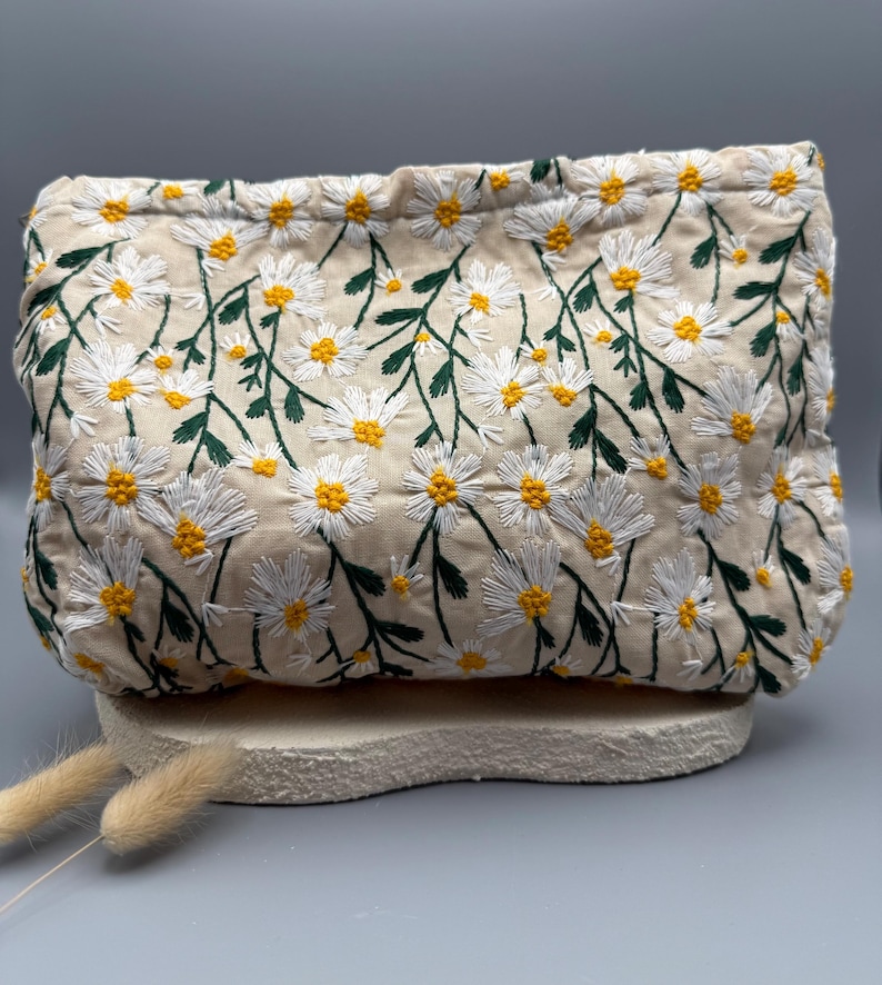 May include: A beige clutch with an embroidered daisy pattern. The daisies have white petals and yellow centers, with green stems and leaves. The clutch is rectangular and sits on a light-colored, textured surface.