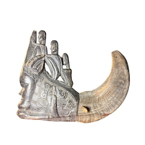 Puede incluir: Una escultura de piedra tallada en gris oscuro que representa a dos figuras montadas a caballo, con intrincados patrones de hojas en la silla. Una regla colocada en la base muestra que el objeto mide aproximadamente 23 cm de largo.