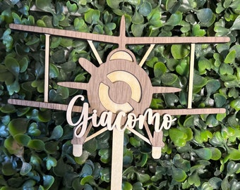 May include: Wooden airplane cake topper with the name Giacomo written in cursive. The airplane is brown and has a light brown propeller. The topper is on a stick.