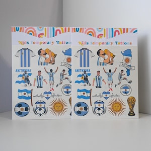 May include: Two sheets of temporary tattoos for kids. Each sheet features images of soccer players, jerseys, flags, and a trophy. The tattoos are in the colors of light blue and white, with a sun and soccer ball design. The text "Kids temporary Tattoos" is printed at the top.