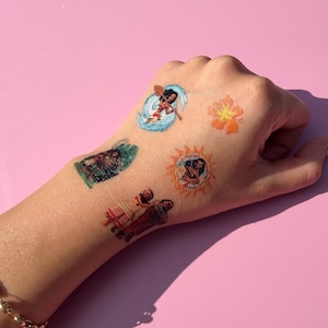May include: A hand adorned with several colorful temporary tattoos. The designs include a surfer, a flower, a sun with a face, and figures in traditional clothing. The tattoos are applied on a light skin tone against a pink background.