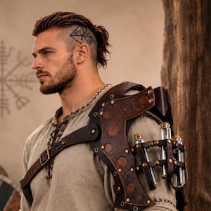 May include: Brown leather shoulder armor featuring intricate embossed circular patterns, metallic rivets, and a harness system. The armor includes a built-in rack holding three clear glass vials with cork stoppers. Text reads: HiiFeuer Medieval Alchemy Shoulder Armor.