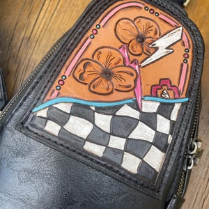 May include: Black leather crossbody bag with a decorative, embossed design. The design features orange flowers, a white lightning bolt, and a checkered pattern. The bag has a zipper closure and an adjustable strap.
