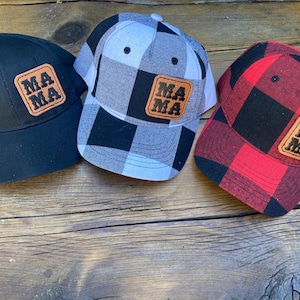Puede incluir: Tres gorras de béisbol con estampado de cuadros de búfalo en blanco y negro, cuadros de búfalo en rojo y negro y tela negra sólida. Cada gorra tiene un parche de cuero marrón con la palabra "MAMA" en letras negras.