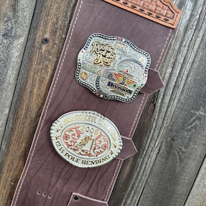 Op de afbeelding: Een bruine leren displayriem met een bovenkant in mandvlechtpatroon. Twee zilverkleurige riemgespen zijn bevestigd: een rechthoekige 2007 Rookie of the Year Utah Wrangler gesp en een ovale 2008 Reserve Champion Pole Bending gesp.