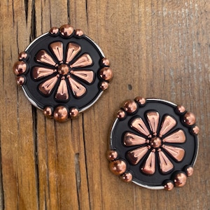 May include: Two circular concho buttons featuring a matte black base with a central copper-toned flower design. The floral motif consists of eight petals surrounding a small centre sphere, accented by small copper spheres along the outer edge.