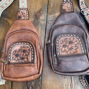 Puede incluir: Dos bandoleras de cuero marrón con diseños de girasoles. Las bolsas tienen un bolsillo frontal con cremallera y correas ajustables con detalles estampados. Una bolsa es de color marrón claro, la otra es de color marrón oscuro.