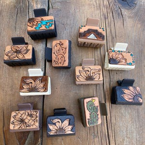 Puede incluir: Una colección de 10 pinzas para el pelo con varios diseños. Las pinzas están hechas de cuero y presentan diferentes patrones, incluyendo girasoles, cactus y diseños geométricos. Las pinzas son todas negras, marrones o blancas.