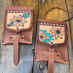 May include: Two brown leather pouches featuring a flap with hand-tooled floral designs. The left pouch displays a single flower with a turquoise arrow, while the right pouch shows two flowers with a turquoise zigzag pattern. Both have leather straps.