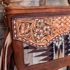 May include: A brown leather handbag with fringe details. The bag has a patterned fabric lining and intricate floral tooling on the front flap.