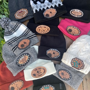 May include: A selection of beanies in shades of black, grey, and rust. Each beanie has a leather patch with designs such as sunflowers, flowers, and a cow. Some beanies have pom-poms. The beanies are arranged on a wooden surface.