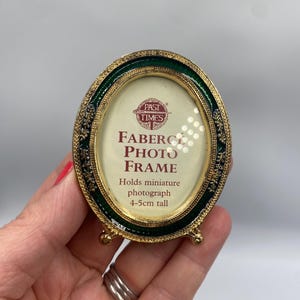 May include: Oval Faberge photo frame with a green enamel border and gold accents. The frame holds a miniature photograph, 4-5cm tall. The frame has a gold base with small feet and the text "PAST TIMES" and "FABERGE PHOTO FRAME".