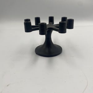 May include: A black candelabra designed to hold seven candles. It features a central stem and a circular base. Seven cylindrical holders extend outwards from a central point. The design is simple and modern, suitable for various settings.