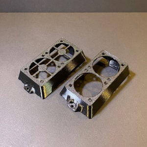 May include: Two black, rectangular 3D-printed components with circular and geometric cutouts. Each piece has a textured surface and a small, protruding feature on one side. The components are designed to fit together.