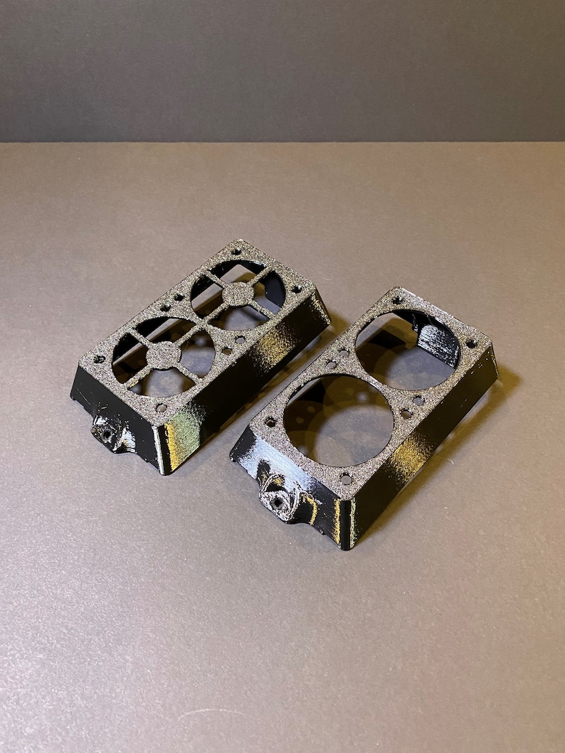 May include: Two black, rectangular 3D-printed components with circular and geometric cutouts. Each piece has a textured surface and a small, protruding feature on one side. The components are designed to fit together.