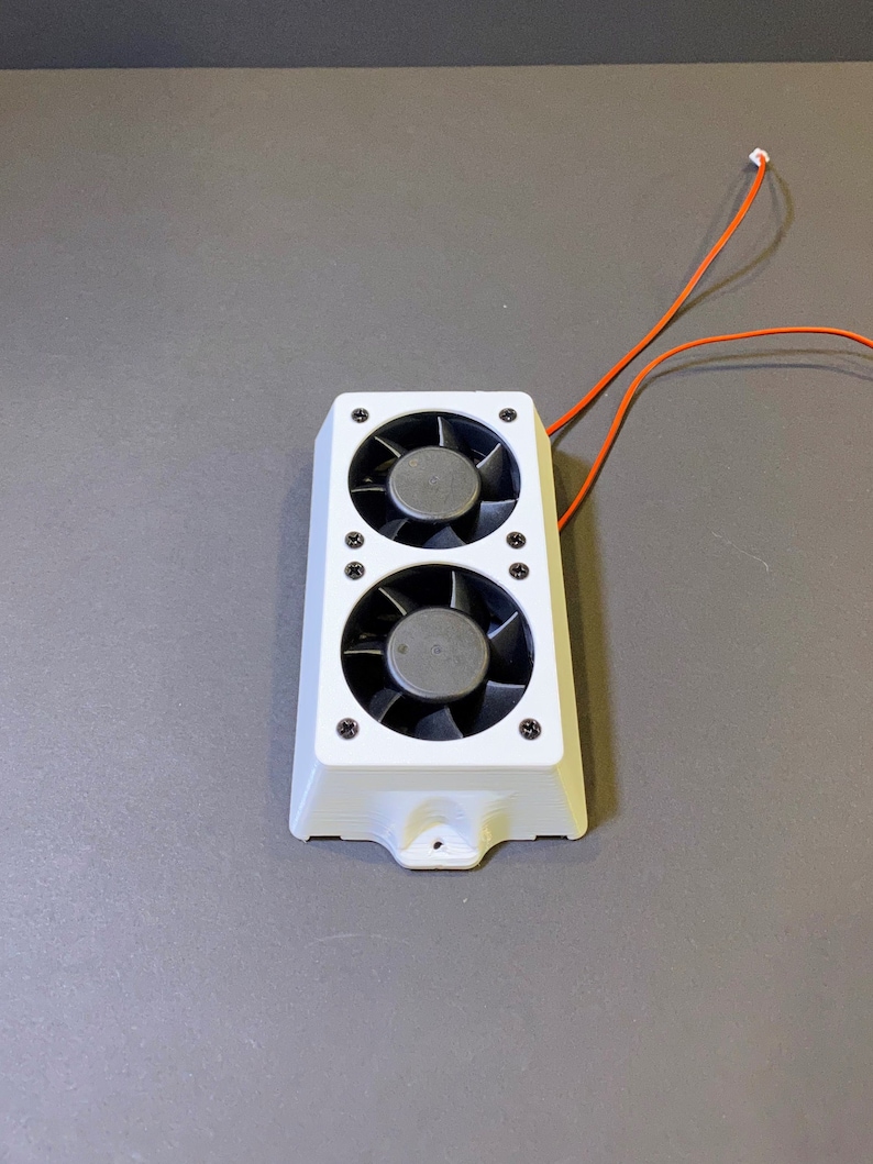 May include: A white electronic component featuring two black fans. The component has a rectangular shape with rounded edges and is secured with black screws. Orange wires extend from the top.