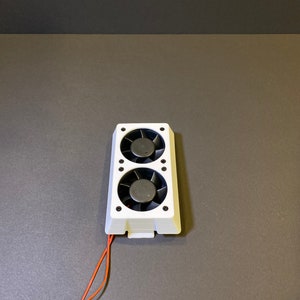 May include: A white electronic component featuring two black fans. The fans are housed in a rectangular frame with black screws. Orange wires extend from the bottom of the component.
