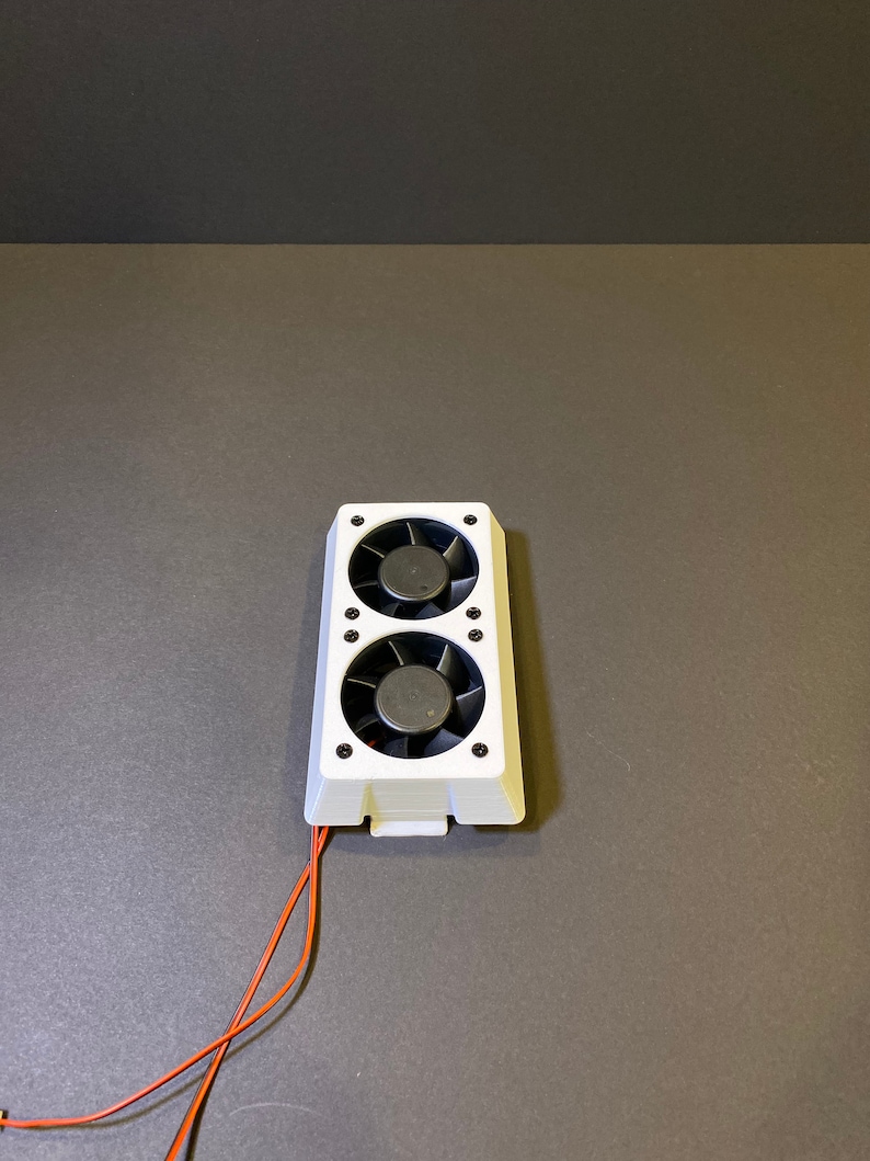 May include: A white electronic component featuring two black fans. The fans are housed in a rectangular frame with black screws. Orange wires extend from the bottom of the component.