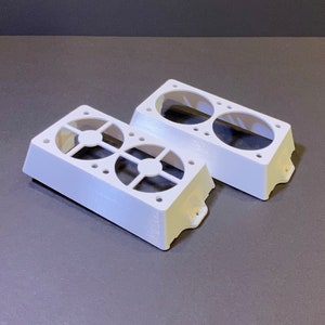May include: Two white, rectangular fan housings with circular openings and a grid-like design. Each housing has two large circular openings and several smaller holes. The housings are made of a solid, opaque material.