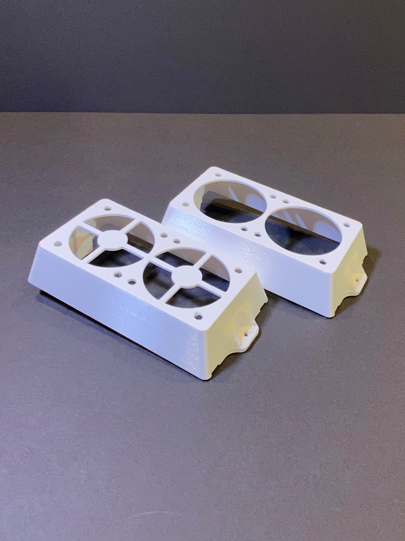 May include: Two white, rectangular fan housings with circular openings and a grid-like design. Each housing has two large circular openings and several smaller holes. The housings are made of a solid, opaque material.