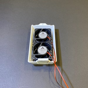 May include: A white rectangular enclosure containing two black brushless DC fans. Each fan has a central label and black blades. Orange and black wires extend from the enclosure.