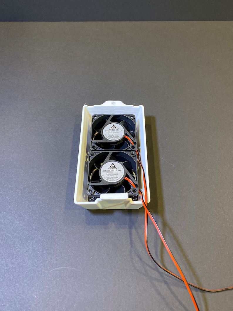 May include: A white rectangular enclosure containing two black brushless DC fans. Each fan has a central label and black blades. Orange and black wires extend from the enclosure.
