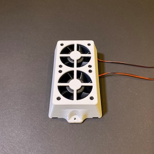 May include: A white rectangular electronic component with two black fans. The component has a series of small black screws and two wires extending from the side. The fans are visible through a grid-like design.