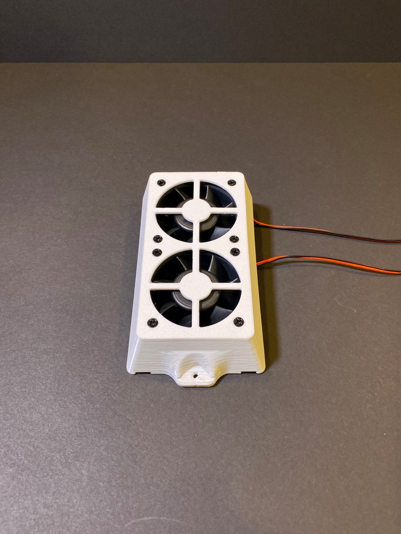 May include: A white rectangular electronic component with two black fans. The component has a series of small black screws and two wires extending from the side. The fans are visible through a grid-like design.