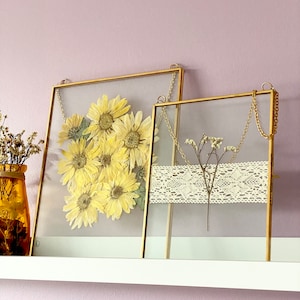 May include: Two gold framed glass picture frames with hanging chains. The frame on the left contains pressed yellow sunflowers. The frame on the right contains a small dried flower arrangement with a white lace ribbon.