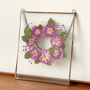 May include: A square glass frame with a brass chain hanging from the top. Inside the frame is a wreath of dried purple flowers and small white flowers.