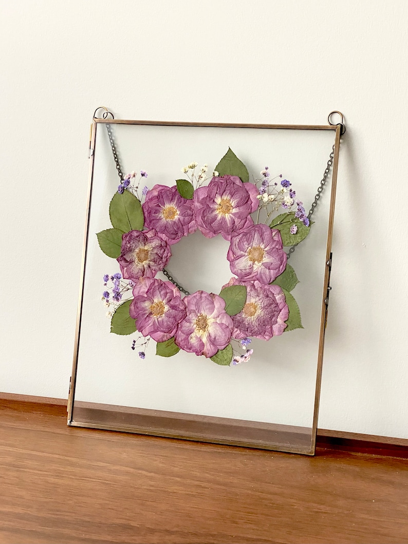 May include: A square glass frame with a brass chain hanging from the top. Inside the frame is a wreath of dried purple flowers and small white flowers.