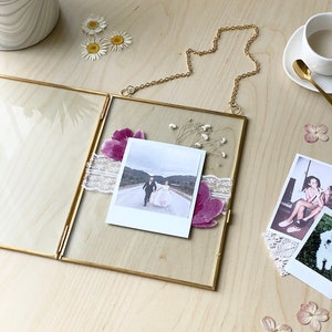 May include: A gold-toned metal frame with a glass insert, holding a Polaroid photo of a couple walking down a road. The frame is hanging from a gold chain and has dried flowers and lace accents.