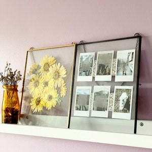 May include: Two glass picture frames hanging on a white shelf against a light pink wall. The frame on the left has a gold metal frame and contains pressed dried yellow flowers. The frame on the right has a black metal frame and contains six Polaroid photos.