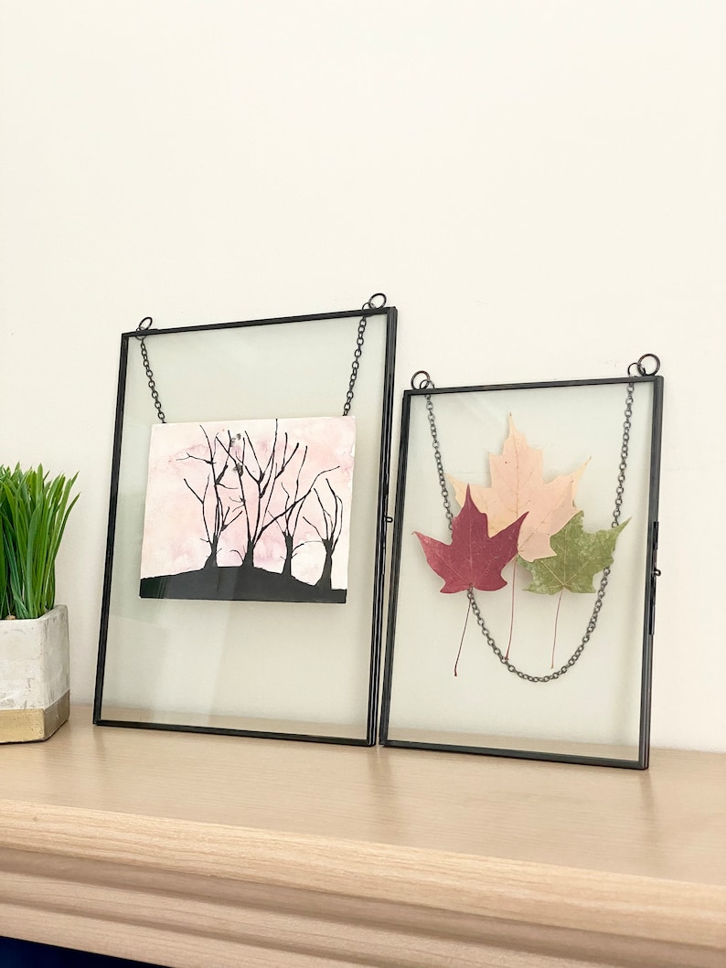 May include: Two black metal picture frames with glass fronts hanging on a chain. The frame on the left contains a black and white drawing of trees against a pink background. The frame on the right contains three pressed leaves, one red, one pink, and one green.