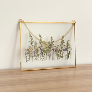 May include: A gold-toned frame with a glass front displays a pressed flower arrangement. The flowers are a mix of white, purple, and green, and are arranged in a horizontal line across the frame.