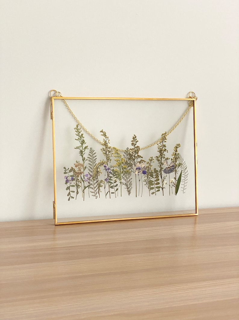May include: A gold-toned frame with a glass front displays a pressed flower arrangement. The flowers are a mix of white, purple, and green, and are arranged in a horizontal line across the frame.