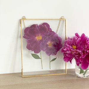 May include: A gold-toned frame with a glass front displays two pressed purple flowers. The flowers are arranged in a simple, elegant design.