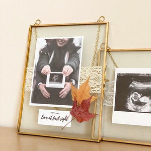 May include: Two gold picture frames with glass fronts, hanging on a wall. The frames contain black and white ultrasound images and a photo of a couple holding a small ultrasound image. The frames are decorated with lace and fall leaves. The text "OCT 2021 love at first sight" is written on a white card in the bottom right corner of the left frame.