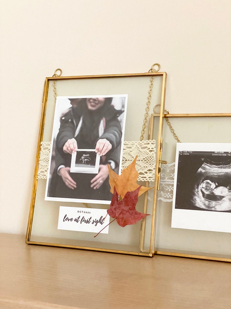 May include: Two gold picture frames with glass fronts, hanging on a wall. The frames contain black and white ultrasound images and a photo of a couple holding a small ultrasound image. The frames are decorated with lace and fall leaves. The text "OCT 2021 love at first sight" is written on a white card in the bottom right corner of the left frame.