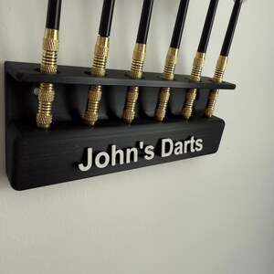 Op de afbeelding: Een zwarte, aan de muur gemonteerde dart houder met acht slots voor darts. De houder heeft de naam "John's Darts" op de voorkant.