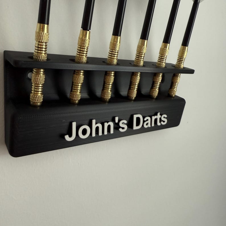 Op de afbeelding: Een zwarte, aan de muur gemonteerde dart houder met acht slots voor darts. De houder heeft de naam "John's Darts" op de voorkant.