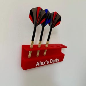 Op de afbeelding: Rode plastic dart houder met zes slots voor darts. Drie darts zitten in de houder. De darts zijn zwart met rode, blauwe en grijze veren. De houder heeft de tekst "Alex's Darts" op de voorkant.