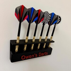 Op de afbeelding: Een zwarte dart houder met een rode inscriptie die "Owen's Darts" luidt. De houder heeft zes vakken en is gevuld met darts met zwarte shafts, gouden tips en kleurrijke flighted tails.