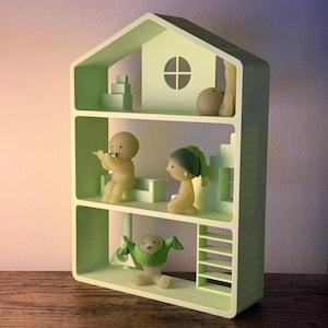May include: A light green three-storey dollhouse shelf featuring a gabled roof and a circular window. Inside, there are several small, smooth, translucent figurines, including one playing a flute, one wearing a green headpiece, and one in a green hood.