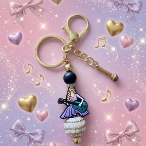 May include: A gold-colored keychain with a charm featuring a woman playing a guitar. The charm is adorned with a black bead, a white and gold beaded section, and a miniature microphone. The background includes bows, hearts, and musical notes.