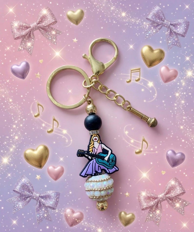 May include: A gold-colored keychain with a charm featuring a woman playing a guitar. The charm is adorned with a black bead, a white and gold beaded section, and a miniature microphone. The background includes bows, hearts, and musical notes.